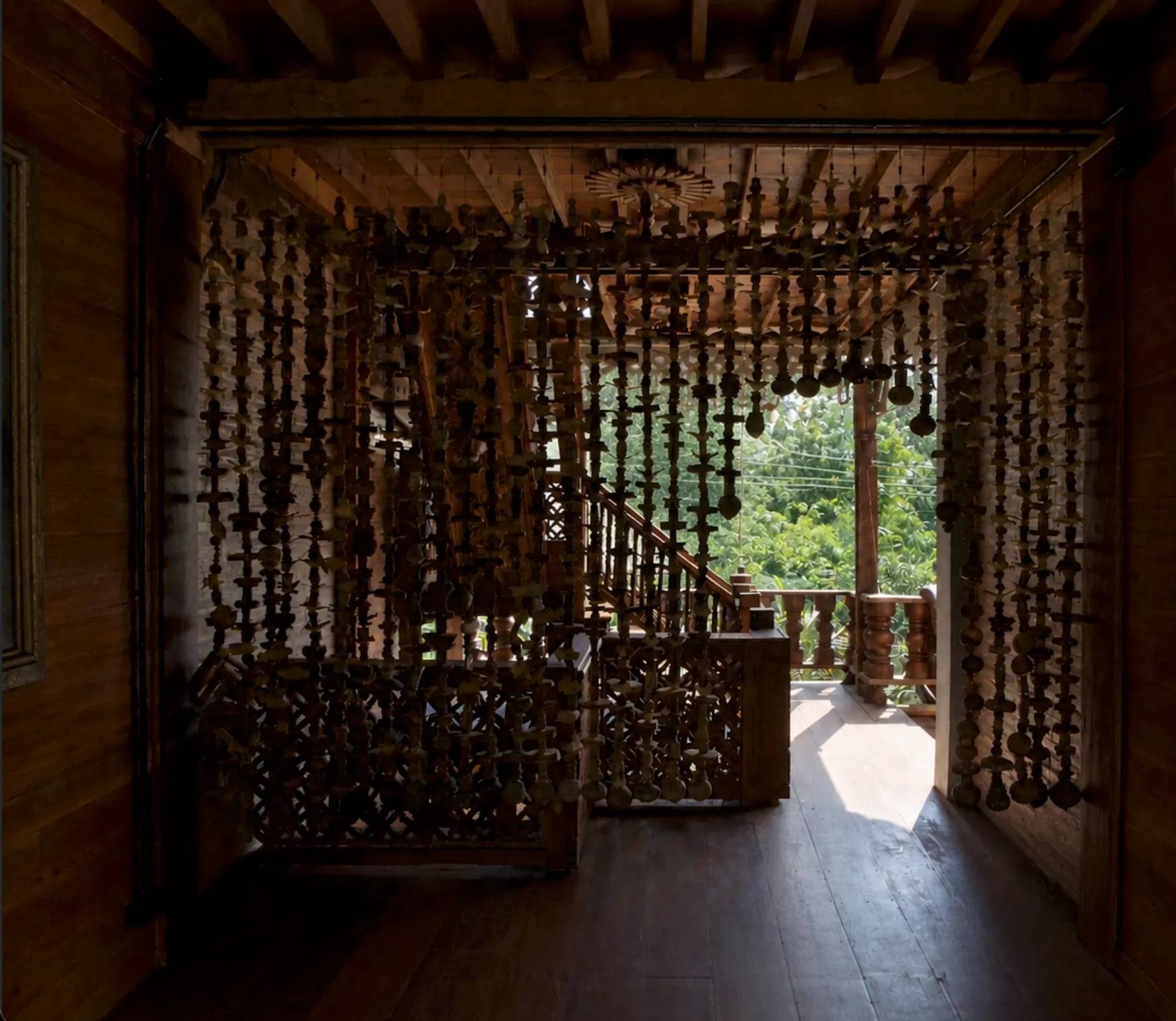 Interior walkway with a hanging curtain of carved beads, a carved staircase and a sunlit teak veranda beyond