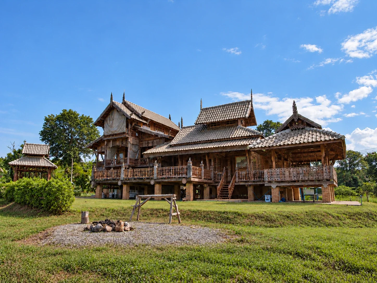 The whole traditional northern Thai teak house, with tiered tile roofs and carved gable finials, seen across the lawn under a blue sky